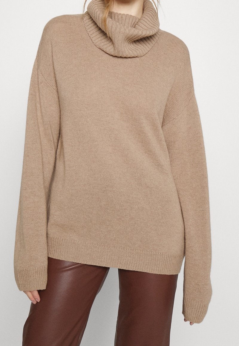 Beige oversized trui met een geribbelde col, textuur breiwerk en lange mouwen; gecombineerd met bruine faux leren broek.