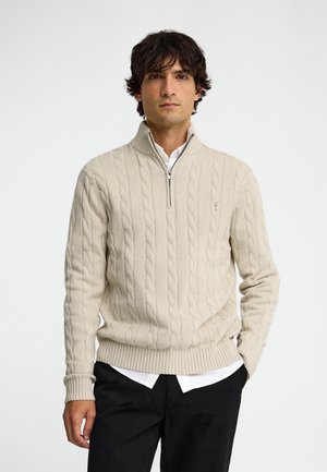 Mann trägt einen beigen Kabelstrick-Viertel-Reißverschluss-Pullover über einem weißen Hemd und schwarzen Hosen, steht vor einem schlichten hellen Hintergrund.