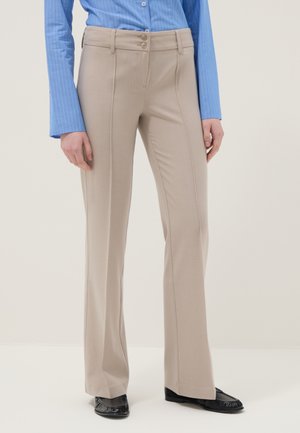 Donna che indossa pantaloni beige su misura a gamba dritta, camicia blu con bottoni e mocassini neri, in piedi contro uno sfondo chiaro e semplice.