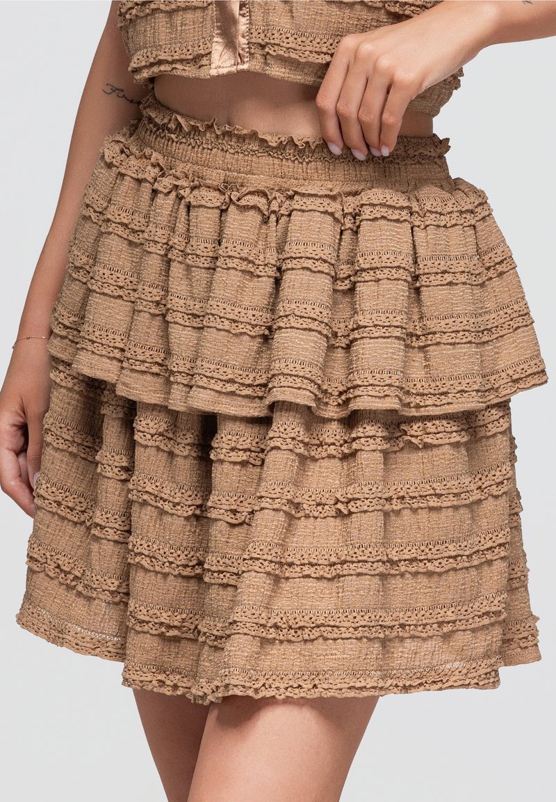 Jupe beige à volants avec tissu texturé et bordure en dentelle, portée à la taille avec le ventre visible et une main reposant sur la hanche.