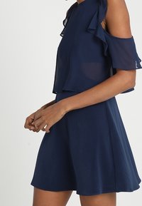 Marinblå klänning med off-shoulder volangärmar, lätt tyg och en flytande kjol som vidgar sig vid nederkanten.