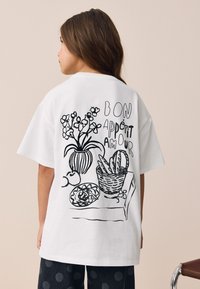 Camiseta blanca de algodón con dibujos en línea negra de un jarrón con flores, frutas y una cesta, con el texto "BON APPÉTIT AMOUR".