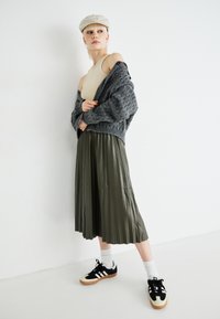 Cardigan de malha cinza sobre um top de tanque creme, saia midi plissada verde-azeitona e ténis pretos com detalhes brancos e solas de borracha.