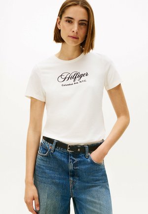 T-shirt blanc à manches courtes arborant un logo script noir "Hilfiger" avec des détails de ville. Associé à un jean en denim bleu et une ceinture noire.