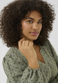 Blouse en tissu texturé vert clair avec des pois noirs, dotée de manches ballon et d'un décolleté en V. Le teint est chaud, avec un maquillage naturel.