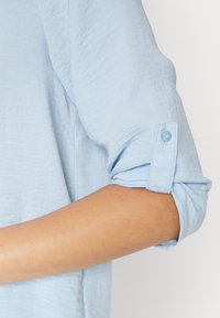 Manica arrotolata di una camicia leggera blu strutturata fissata con una cinghia a bottone, che mostra un avambraccio nudo su uno sfondo bianco.