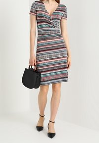 Femme portant une robe rayée à manches courtes et longueur genou, tenant un sac à main noir, assortie à des talons noirs pointus avec brides à la cheville.