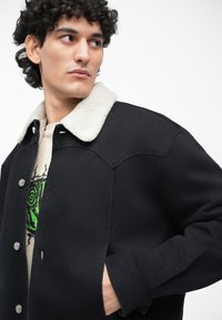 Chaqueta de mezclilla negra con collar de borrego, presenta herrajes de botón plateado, bolsillos laterales y un corte relajado. Decorada con una camiseta gráfica verde debajo.