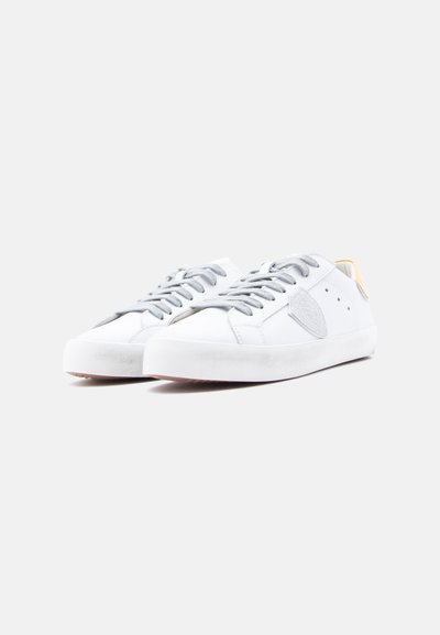 Λευκά πάνινα αθλητικά παπούτσια με γκρι κορδόνια, υφασματικό γκρι τμήμα στο πλάι και κίτρινη ετικέτα στη φτέρνα. Επίπεδη σόλα από καουτσούκ.