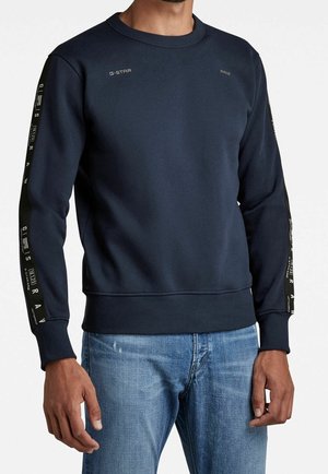Mężczyzna w granatowej bluzie z napisem G-Star Raw na piersi i rękawach, w połączeniu z jasnoniebieskimi dżinsami, na jednolitym białym tle.