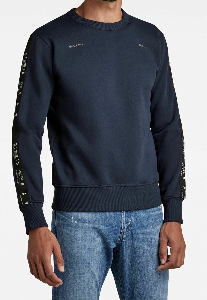 Homme portant un sweat-shirt bleu marine avec le texte G-Star Raw sur la poitrine et les manches, assorti à un jean bleu clair, sur un fond blanc uni.