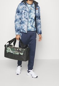 Man som bär en blå och vit mönstrad jacka och byxor, håller en svart och camouflagemönstrad Nike-duffelväska, står mot en enkel ljus bakgrund.