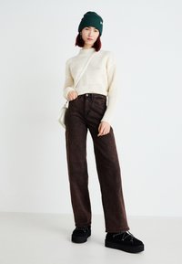 Maglione lavorato a maglia crema con motivi geometrici, design a collo alto, abbinato a jeans a gamba larga marroni scuri e scarpe con plateau nere con accenti bianchi.