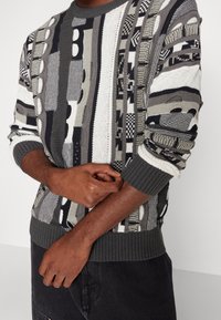 Gebreide trui in gedempte tinten van grijs en wit, met verschillende patronen en texturen. Geribbelde boorden en zoom met contrasterende kleuren.