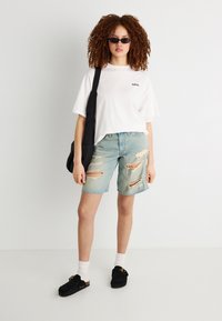 Νέα γυναίκα με σγουρά μαλλιά φοράει γυαλιά ηλίου, λευκό υπερμεγέθες μπλουζάκι, σκισμένο τζιν σορτς, μαύρη τσάντα ώμου, λευκές κάλτσες και μαύρα παπούτσια χωρίς κορδόνια.