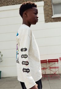 Lysegrå langærmet skjorte med farverige skateboardsgrafikker på forsiden og sort tekst på ærmerne; afslappet pasform, blød tekstur.