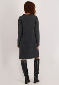 Vrouw met bruin haar in een lange, donkergrijze jurk tot de knie met lange mouwen en zwarte kniehoogte hakken, met de rug naar de camera tegen een effen achtergrond.
