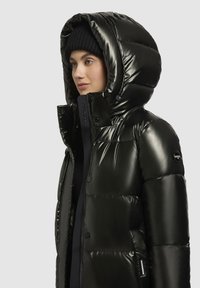 Vadderad svart pufferjacka med hög krage och överdimensionerad huva, med glansig finish, knappar och en logotyp-patch på ärmen.