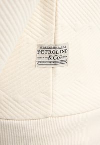 Crèmekleurige stof met diagonale ribbels; bevat een label met zwarte tekst met merk- en registratiegegevens.