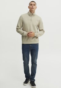Sudadera de cremallera beige con cuello alto, de mangas largas y textura suave, combinada con jeans azul oscuro y zapatillas negras, que presenta un logo sutil en el pecho.