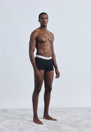 Black boxer briefs with a white branded waistband, displayed on a male model standing on a gray surface against a plain backdrop.
