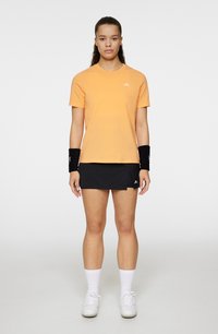 Orange bomullströja med rund hals, i kombination med en svart kjol. Svarta handledsrandsbär och vita sneakers fulländar den atletiska stilen.