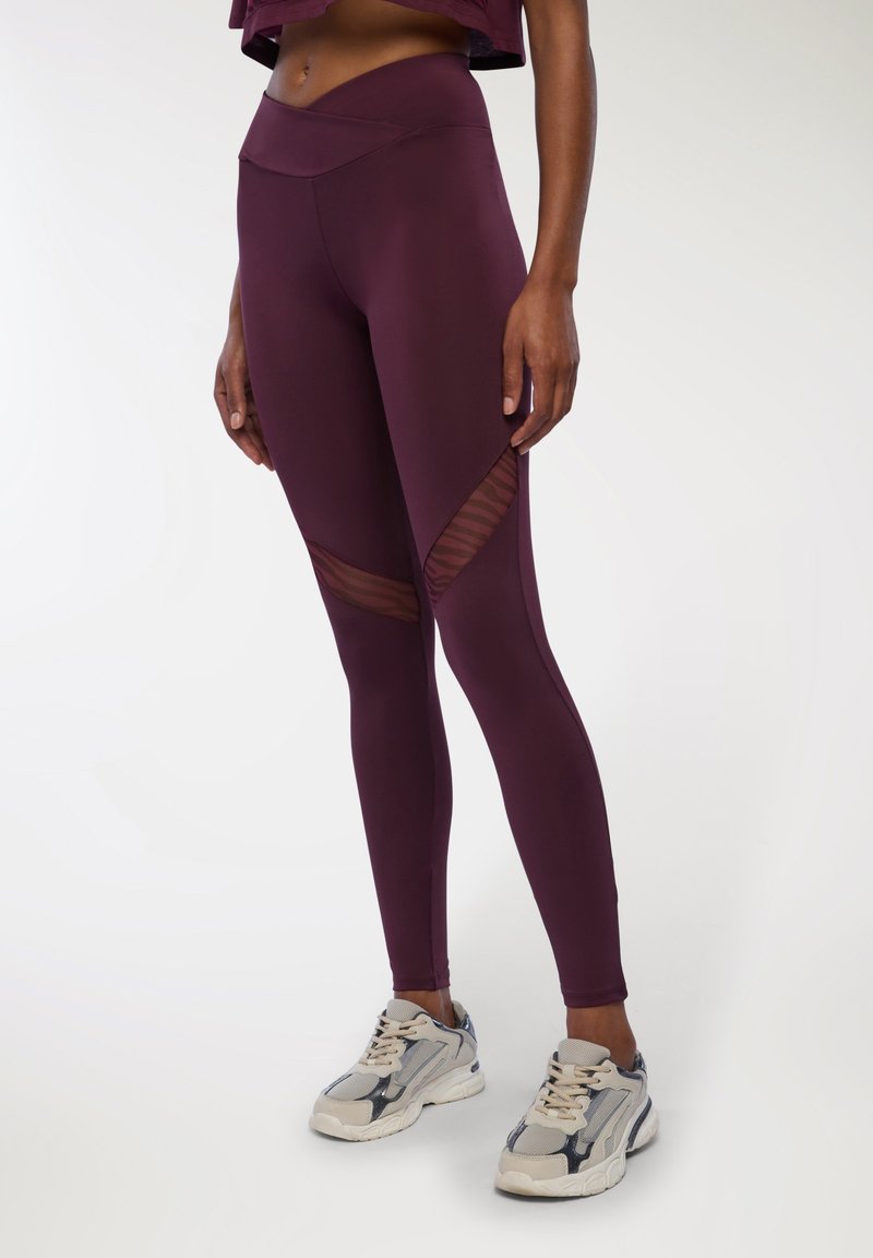 Leggings bordeaux realizzati in tessuto elastico con pannelli in rete e una vita alta, abbinati a scarpe da ginnastica di colore chiaro.