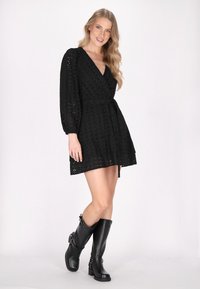 Schwarzes Kleid mit floralem Durchbruchmuster, V-Ausschnitt, langen Puffärmeln, tailliertem Bund und gestuftem Rock. Kombiniert mit knielangen schwarzen Stiefeln.