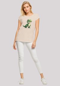 Beige short-sleeve t-shirt featuring a green dinosaur graphic. Paired with white skinny jeans and white sneakers. Plain background.