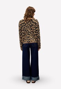 Vrouw met krullend bruin haar, gekleed in een trui met luipaardprint, een wijdvallende donkerblauwe spijkerbroek met omslag en schoenen met luipaardprint, staat met haar rug naar de kijker.