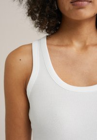 Valge ribane tank top laia rihmaga ja ümarate kaelusjoontega, esitatud mudeli ülaselja peal, omades siledat tekstuuri ja puhtaid jooni.