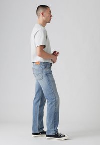Lichtgrijs T-shirt met korte mouwen, gecombineerd met blauwe jeans met rechte pijpen. De jeans hebben klassieke stiksels en een leren label op de achterste tailleband.
