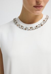 Nahaufnahme eines weißen ärmellosen Oberteils mit einem runden Halsausschnitt, verziert mit abwechselnden braunen und klaren Perlenclusters.