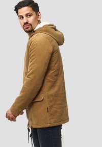 Hombre con una chaqueta de pana color beige con capucha y forro de felpa blanca, mirando hacia un lado contra un fondo gris liso.