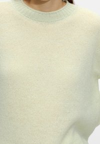 Pull en maille vert clair avec un col côtelé, présentant une texture douce et une coupe ample. Le tissu semble respirant et léger.