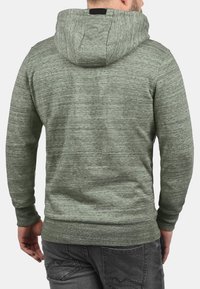 Hombre con una sudadera verde texturizada y jeans gris oscuro, visto de espaldas contra un fondo claro y liso.