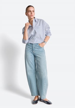 Femme portant une chemise à rayures bleues et blanches à boutons, un jean large bleu clair et des ballerines noires, debout avec une main dans la poche.