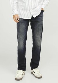 Jeans din denim albastru închis cu o croială dreaptă, prezentând o ușoară decolorare, asortați cu adidași albi și gri. Se poartă o cămașă cu mânecă lungă, cu dungi.