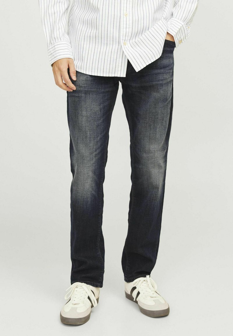 Jeans din denim albastru închis cu o croială dreaptă, prezentând o ușoară decolorare, asortați cu adidași albi și gri. Se poartă o cămașă cu mânecă lungă, cu dungi.