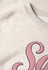 Tissu couleur crème avec encolure côtelée et vue partielle d'une lettre brodée texturée rose et bleue.