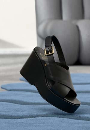 Sandale compensée en cuir noir avec bride à la cheville et boucle dorée reposant sur une surface bleue texturée, avec une chaise grise tapissée en arrière-plan.