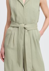Jumpsuit senza maniche di un verde oliva chiaro, con colletto, cintura da annodare in vita e tasche laterali, realizzato in un tessuto texturizzato.