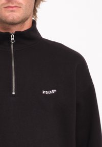 Sudadera de forro polar negra con un collar con cremallera y un pequeño parche blanco con el logotipo en el lado izquierdo del pecho. Textura suave y corte holgado.