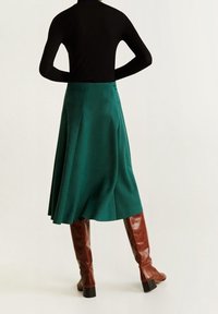 Falda de ante verde con un diseño acampanado, combinada con botas altas de charol marrón. La parte superior es negra y ajustada, en contraste con la falda.