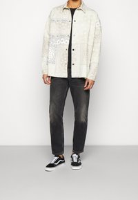 Giacca camicia a fantasia crema e grigia con stampa bandana, t-shirt nera sotto, abbinata a jeans neri sbiaditi e sneakers nere.