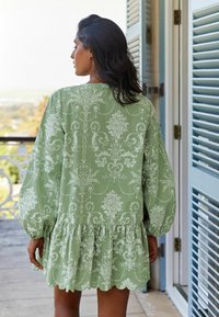 Vihreä kuvioitu mekko, jossa on pitkät kukkakuosiset pallohiha, röyhelöhelma ja kukkakuosit. Pehmeä, teksturoitu kangas, jossa on aaltoileva reunat.