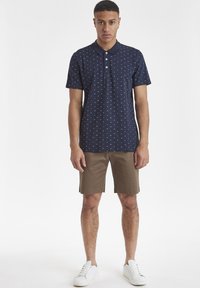 Camisa polo marina con patrones geométricos blancos, mangas cortas y un cuello. Combinada con pantalones cortos marrón claro y zapatillas blancas.