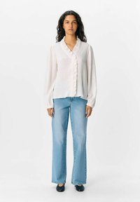 Blusa bianca a maniche lunghe con colletto arricciato e chiusura a bottoni, abbinata a jeans a gamba larga azzurri e scarpe slip-on nere.