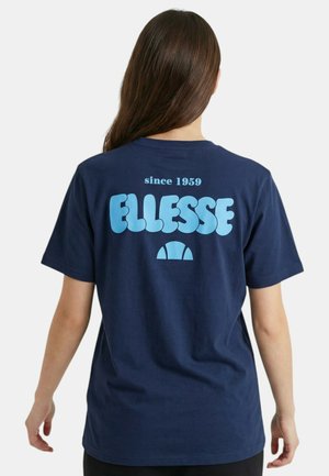 Donna con lunghi capelli castani che indossa una maglietta blu navy con testo e logo "since 1959 ELLESSE" azzurro chiaro sulla schiena.