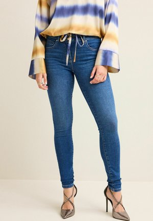 Woman wearing blue skinny jeans, a yellow and purple striped blouse, and metallic high-heeled shoes with ankle straps.